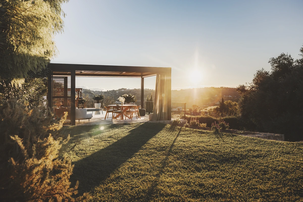 “Pérgola Camargue marrón con paneles laterales deslizantes y cortinas de textil exterior, iluminada por la luz del atardecer para crear un ambiente acogedor y elegante.