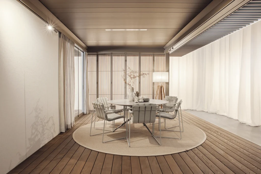 Pérgola Amani Renson beige con paneles de cristal y cortinas exteriores al atardecer